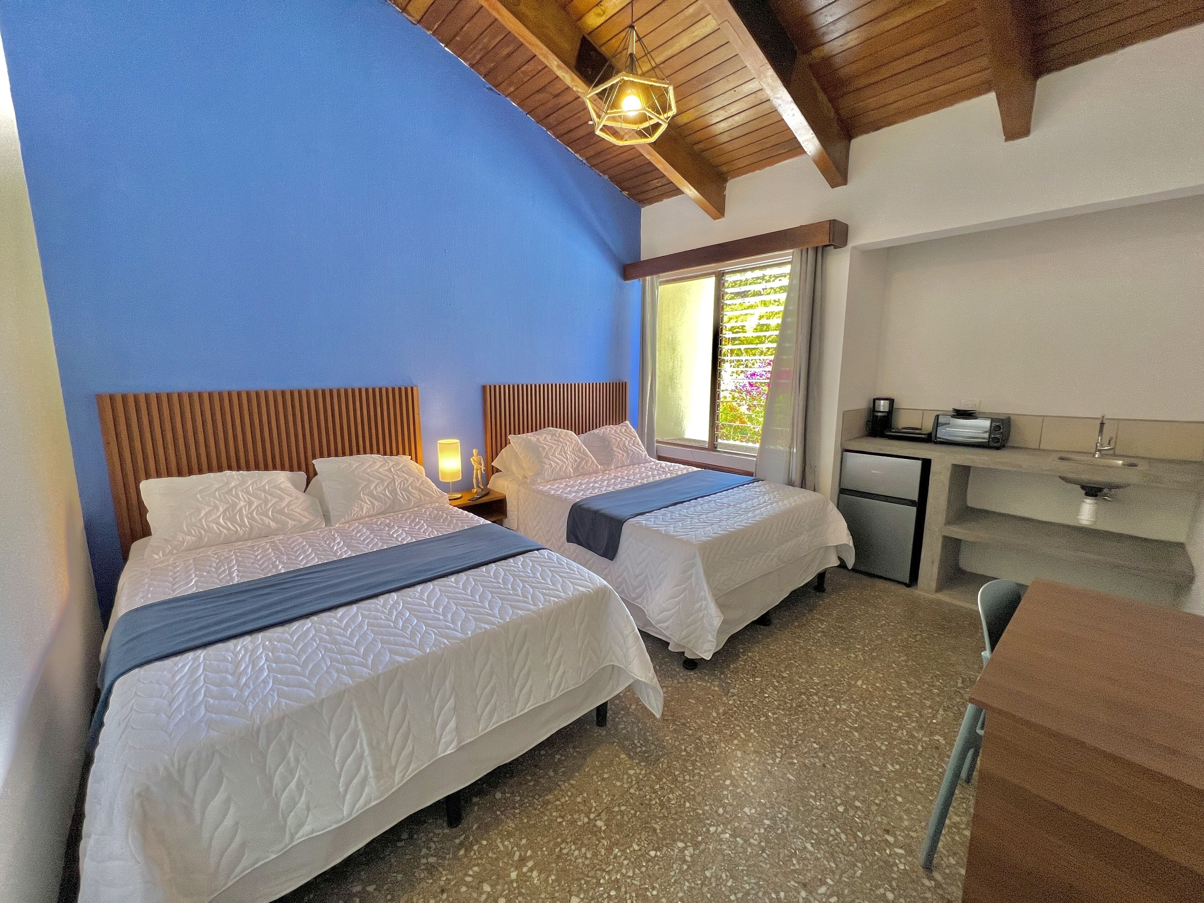 A hotel room with two beds, a small kitchenette, and a window with a view of greenery.