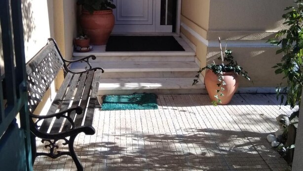 Un banco de madera, una puerta blanca con un panel de vidrio y plantas en macetas sobre un patio con baldosas.