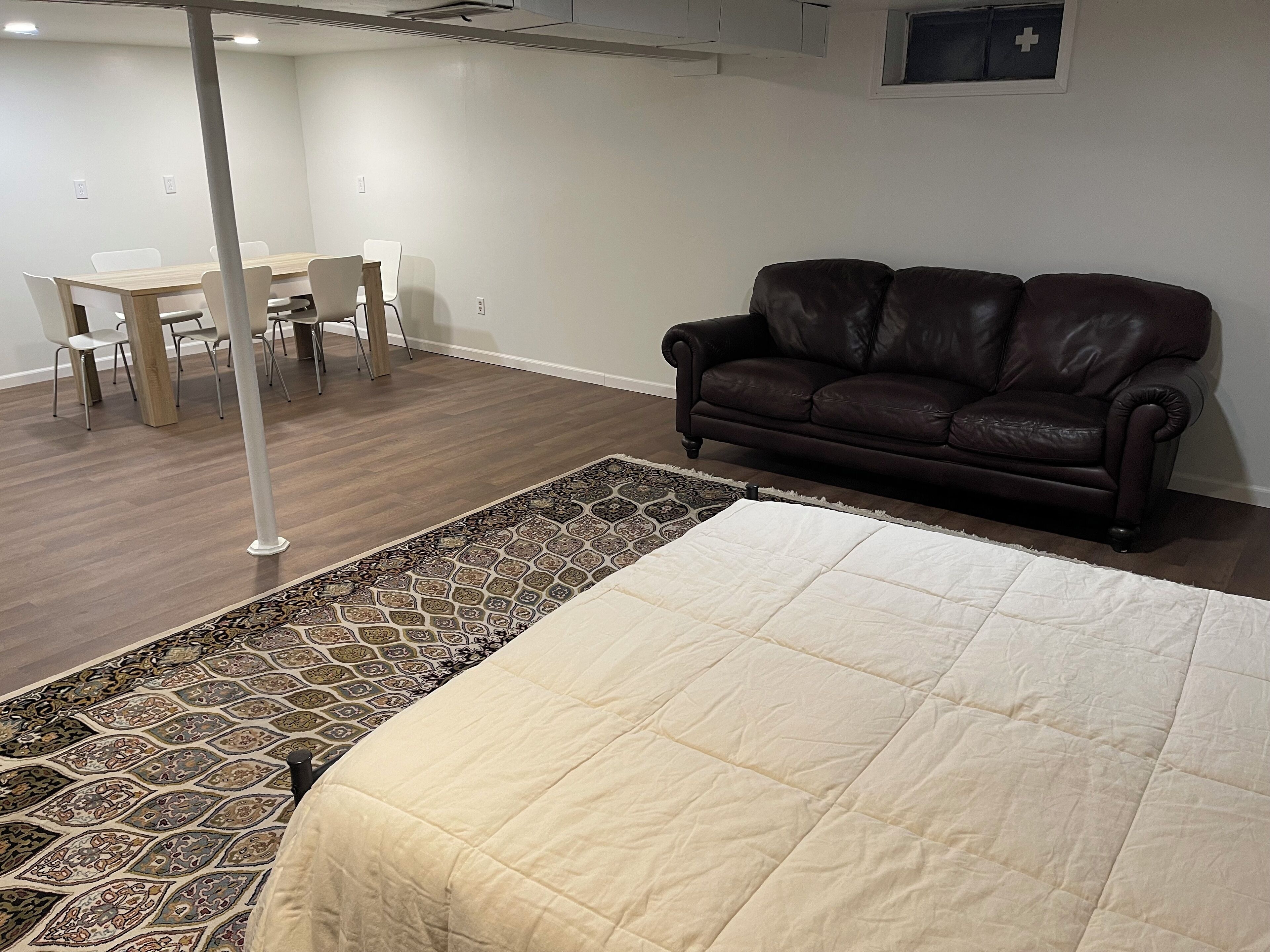 A room with a large bed, a brown leather sofa, a dining table with chairs, and a patterned rug.