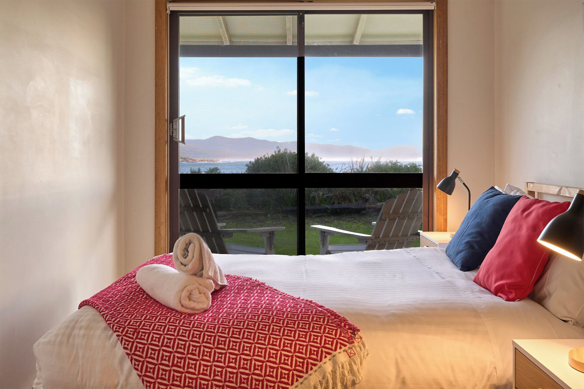 Une chambre avec une grande fenêtre, un lit recouvert d’une couverture à motifs, deux oreillers, une lampe de chevet et une vue sur un jardin et des montagnes.