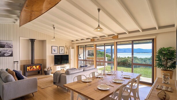 A spacious living area with a fireplace, a dining table set for six, and a large window offering a view of the water. - Waubs Beach Cottage - Bicheno Accommodation (Glamorgan-Spring Bay)