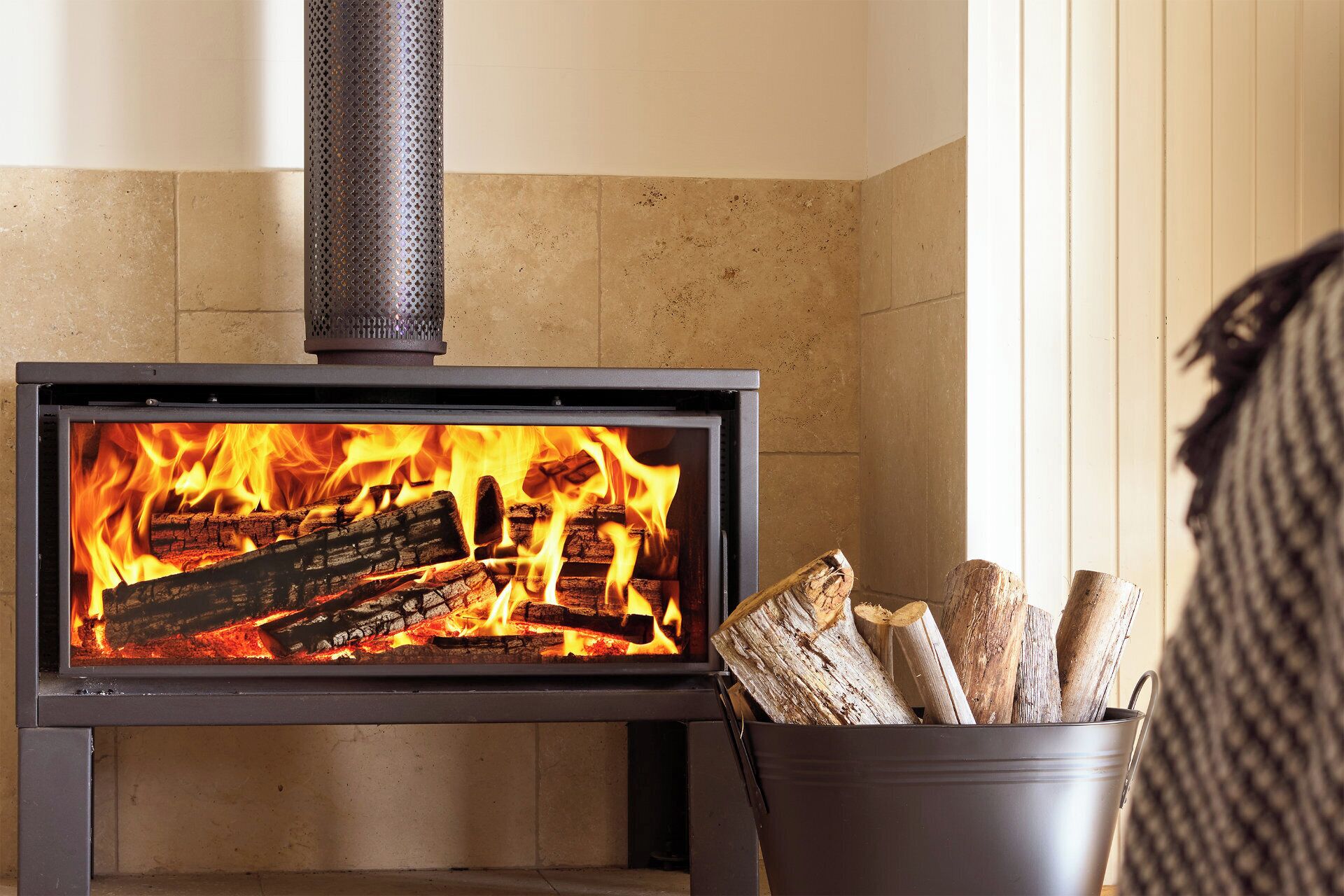 A wood-burning stove with a visible fire, a metal bucket with logs, and a textured wall.