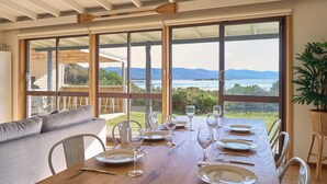 A dining area with a table set for six, large windows offering a view of the sea and mountains, and a cozy seating area with a sofa and chairs. - Waubs Beach Cottage - Bicheno Accommodation (Glamorgan-Spring Bay)