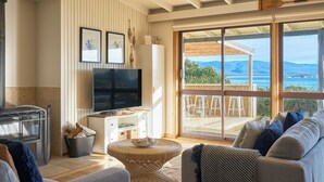 A cozy living room with a large TV, comfortable seating, and a view of the sea. - Waubs Beach Cottage - Bicheno Accommodation (Glamorgan-Spring Bay)