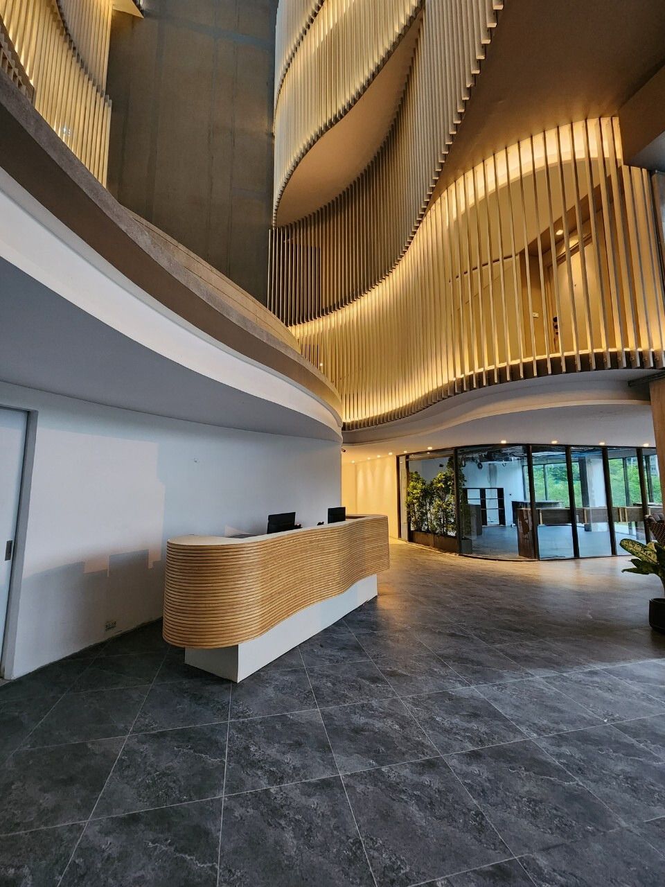 A modern reception area with a curved wooden reception desk, a high ceiling with vertical slats, and a view of the interior through large glass doors.