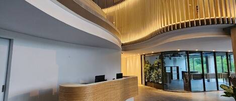 A modern reception area with a curved wooden reception desk, a high ceiling with vertical slats, and a view of the interior through large glass doors.