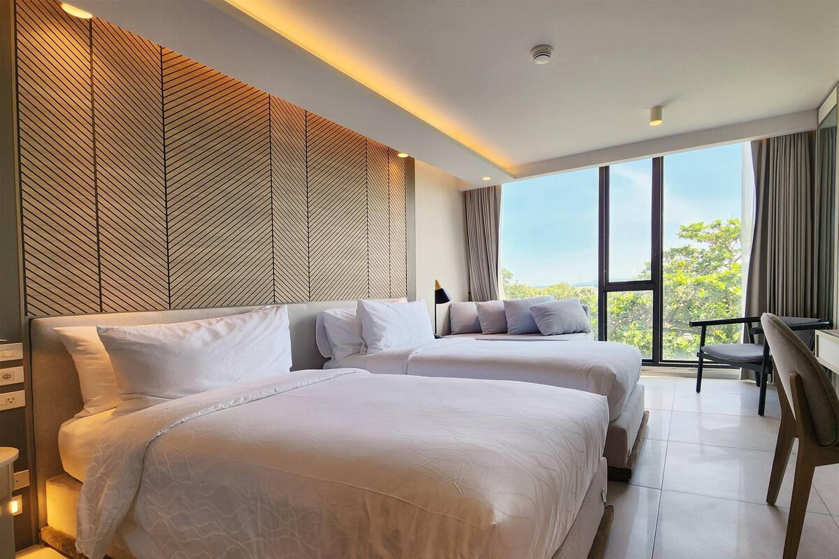 A modern hotel room with two beds, a desk, and a view of the outdoors.