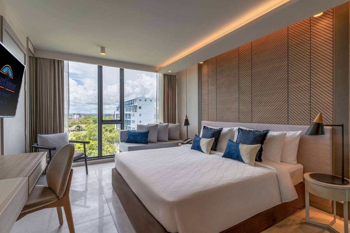 A modern hotel room with a large bed, a desk, and a sofa. There is a view of the city through large windows.