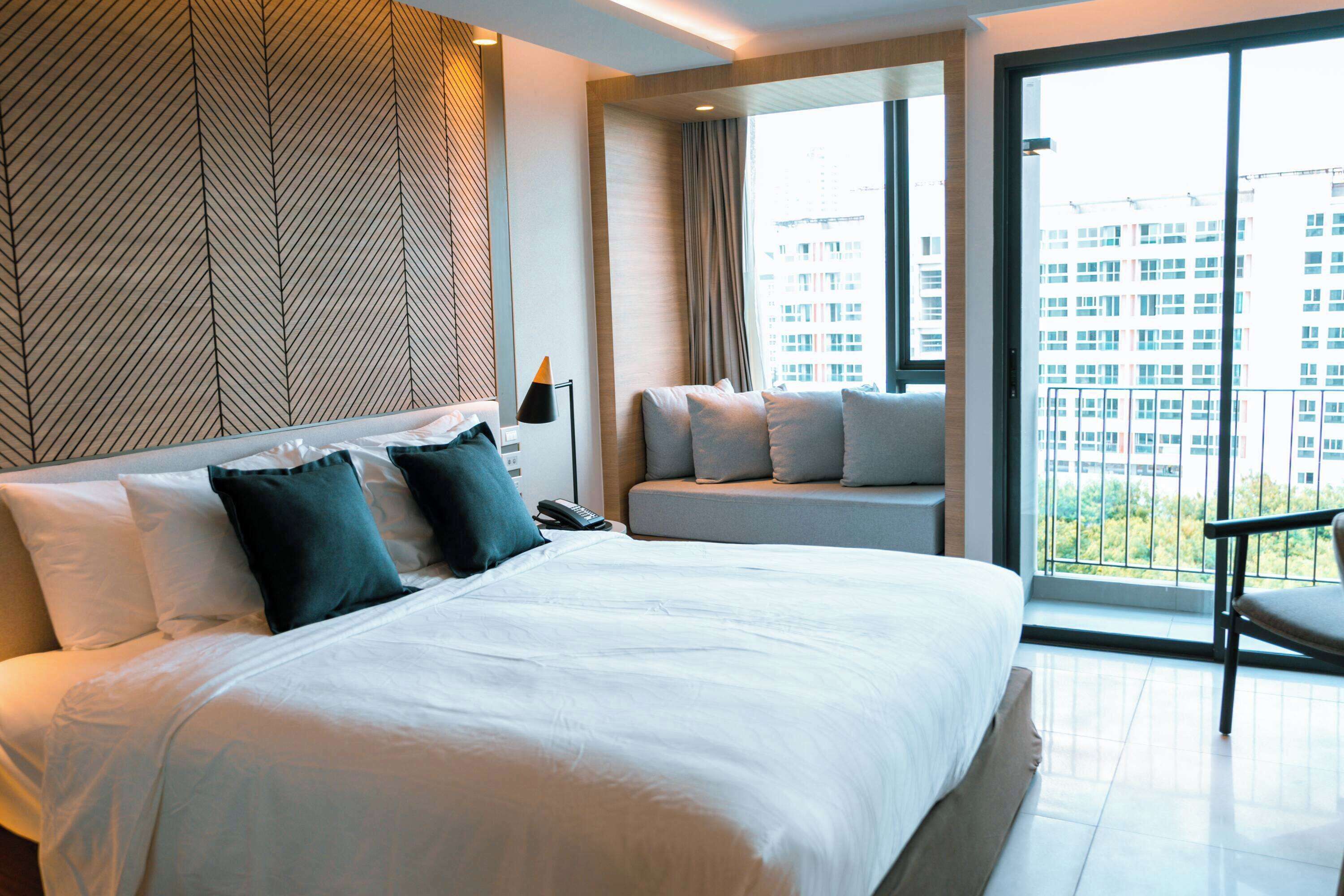 A modern hotel room with a large bed, a sofa, a chair, and a view of the city through large windows.