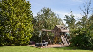 A wooden playhouse with a slide and climbing wall, surrounded by trees and a grassy yard.