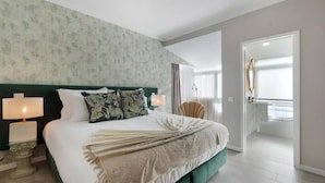 A modern hotel room with a large bed, two bedside lamps, a green headboard, floral pillows, a beige throw blanket, and a view of the bathroom through an open door. - Impeccable 4-Bed House in São Domingos de Rana (Sao Domingos de Rana)