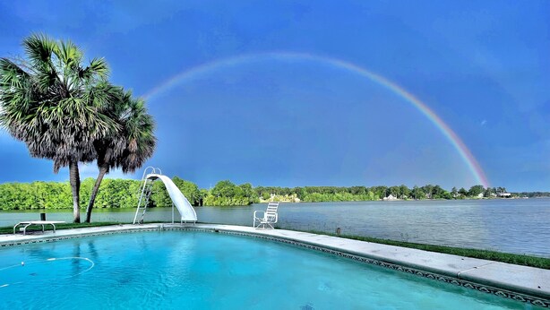 A swimming pool with a slide, palm trees, and a view of a lake with a rainbow.