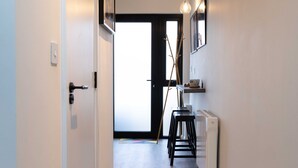 Un couloir avec une porte, un radiateur, un tabouret et une étagère sur laquelle se trouve une tasse.
