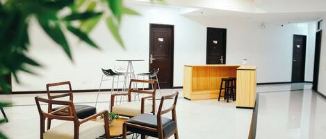 A modern hotel lobby with a reception desk, wooden chairs, a small table with a plant, and a large potted plant in the foreground.