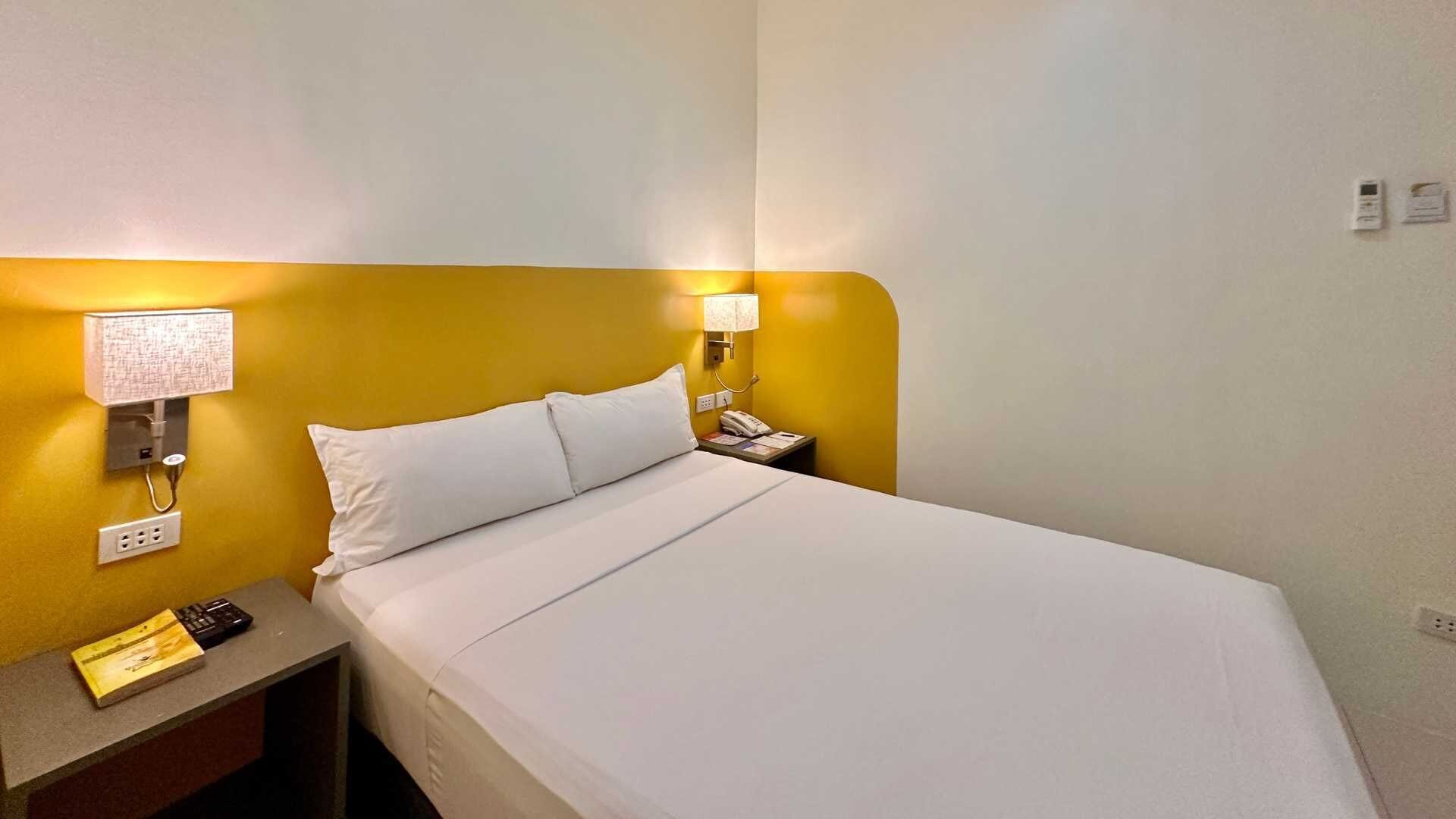 Habitación de hotel con cama, lámpara de noche, teléfono y una mesita con un libro y un control remoto.