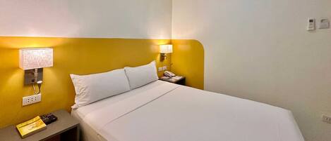 Habitación de hotel con cama, lámpara de noche, teléfono y una mesita con un libro y un control remoto.