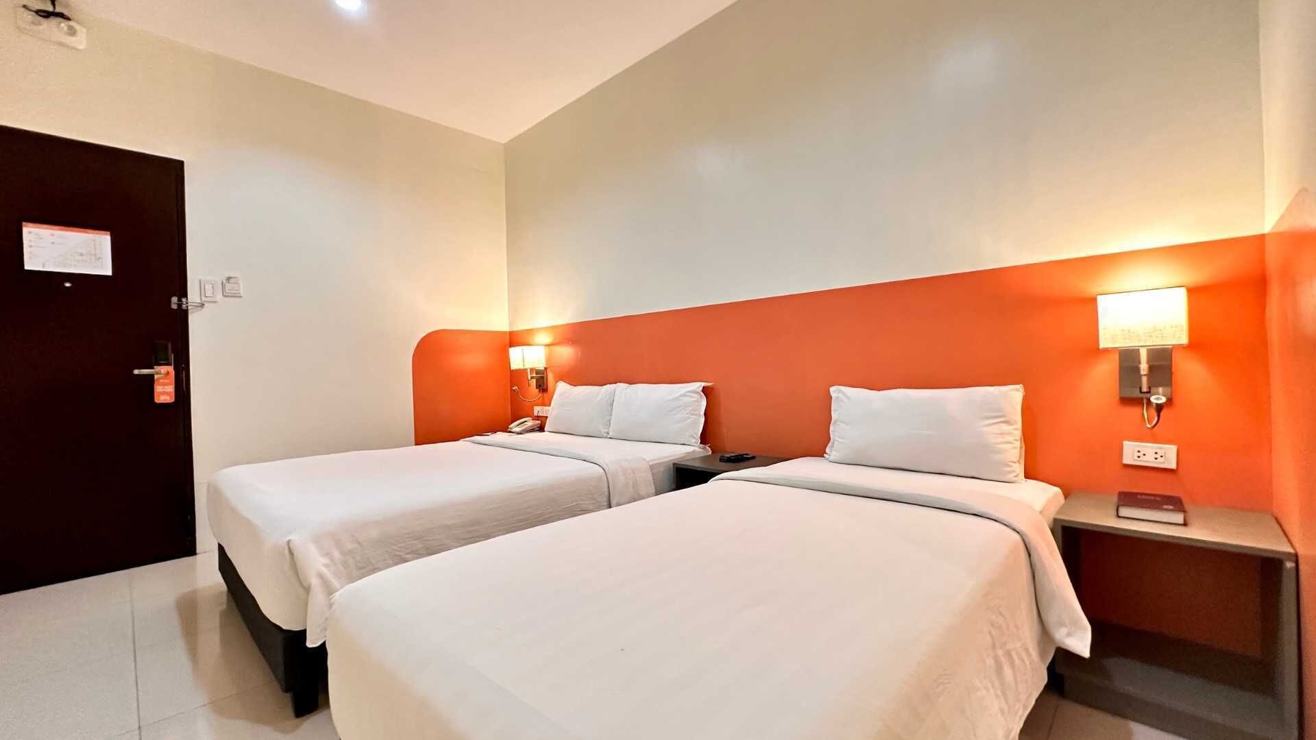 A hotel room with two beds, a door, a nightstand with a lamp, and a wall-mounted light.