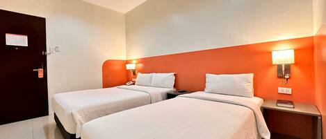 A hotel room with two beds, a door, a nightstand with a lamp, and a wall-mounted light.