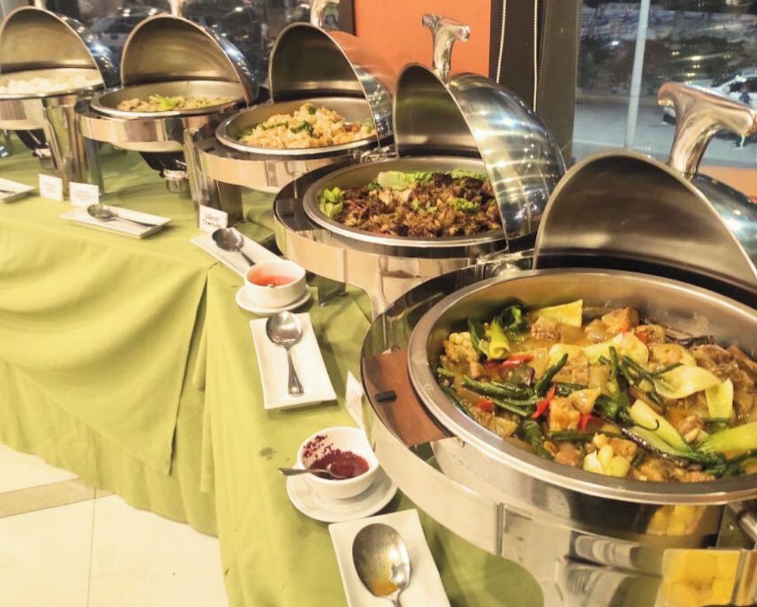 Montaje de bufé con diversos platos en recipientes metálicos, un mantel verde y una cuchara y servilleta sobre la mesa.