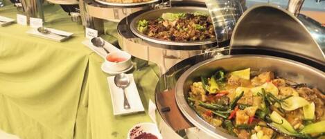 Montaje de bufé con diversos platos en recipientes metálicos, un mantel verde y una cuchara y servilleta sobre la mesa.