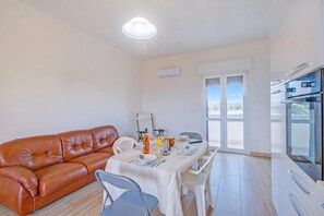 Un salon moderne avec un canapé en cuir marron, une table à manger dressée avec des assiettes, une nappe blanche et un coin cuisine équipé d’un réfrigérateur et d’un four.