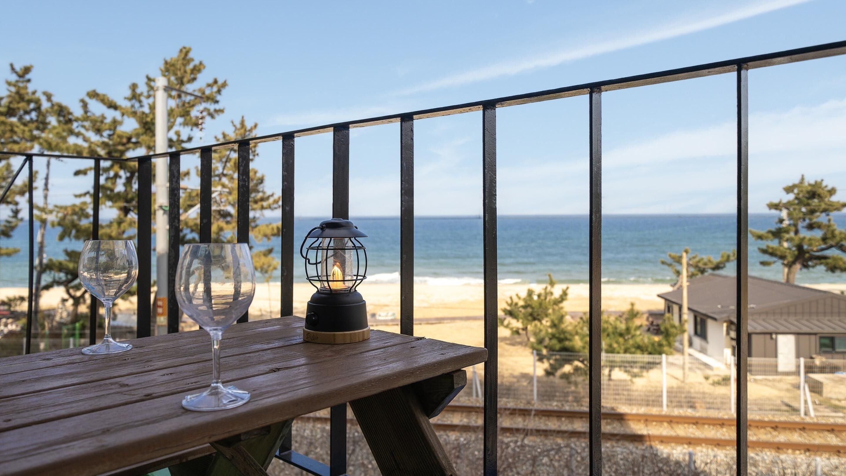 Mesa de madeira com dois copos de vinho e um candeeiro numa varanda com vista para a praia.