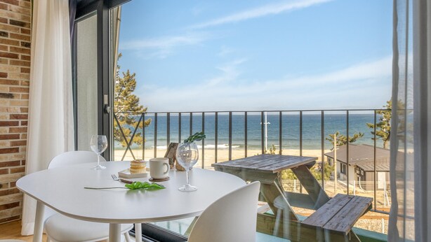 A balcony with a table set for two, overlooking a beach and ocean. - Gangneung Jd38 spa Pension (Gangneung)