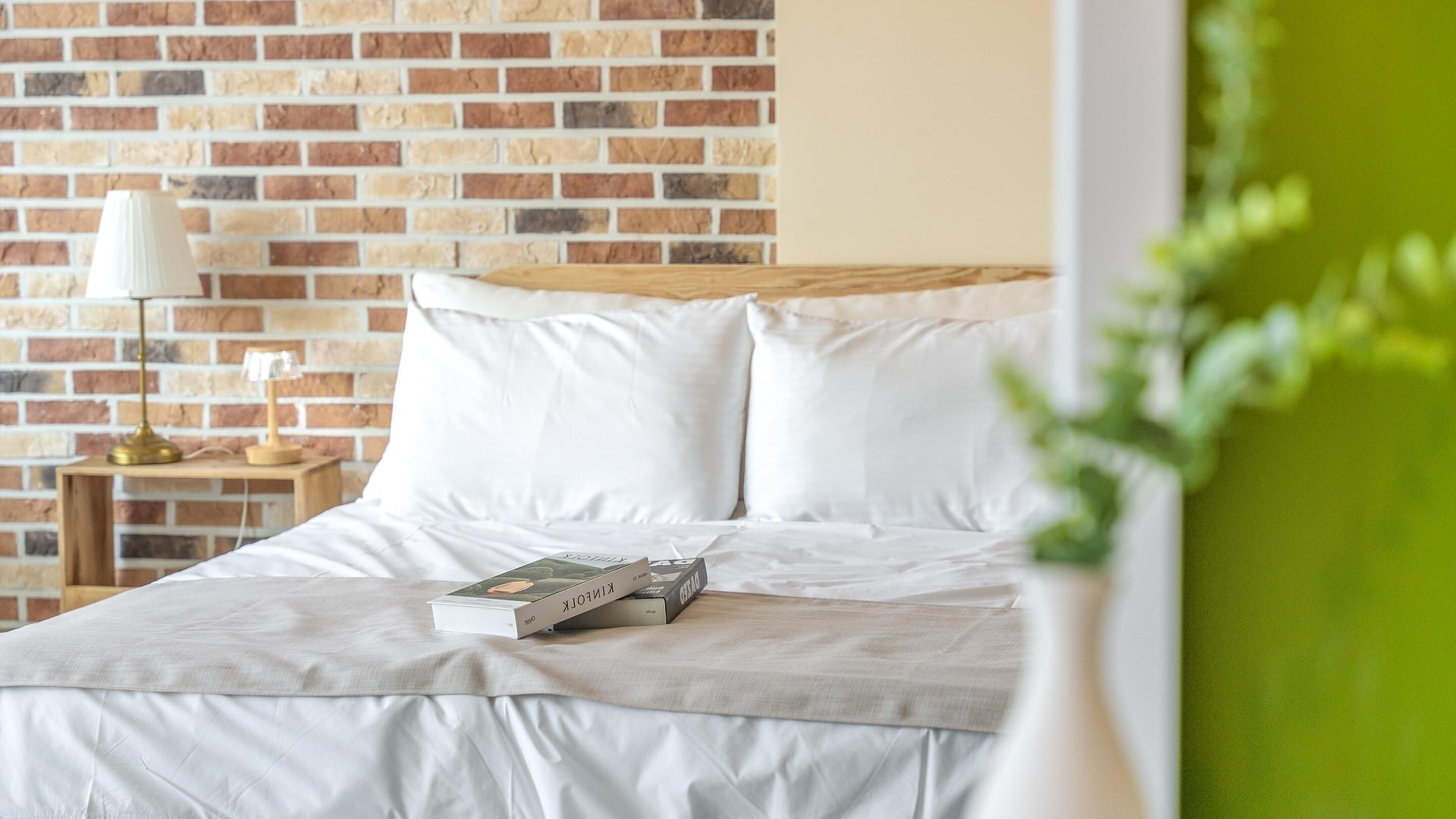 Una cama bien hecha con sábanas blancas, un libro sobre la cama y una pared de ladrillo al fondo.