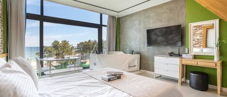 A modern bedroom with a large bed, a desk, a TV, and a balcony with a view of the sea.
