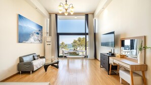 A modern hotel room with a large window, a flat-screen TV, a desk, a sofa, and a painting of a coastal scene. - Gangneung Jd38 spa Pension (Gangneung)