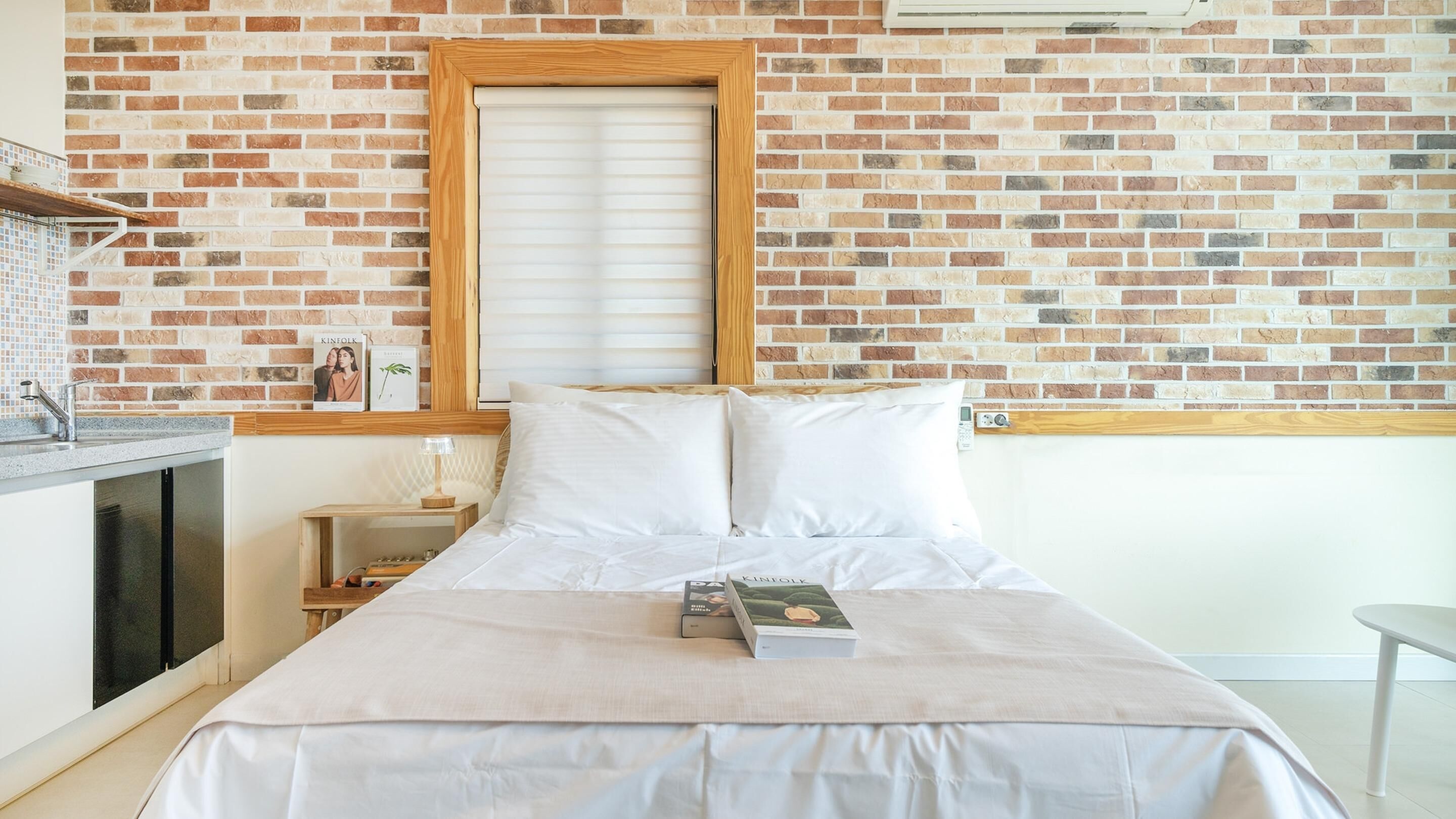 Une chambre avec un mur décoré d’un motif en briques, une fenêtre à cadre en bois, un lit avec une literie blanche, une table de chevet avec une lampe et une petite étagère avec des livres.