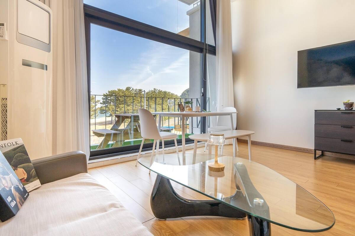 Een moderne woonkamer met een glazen tafel, een bank, een aan de muur bevestigde televisie en een balkon met tuinmeubilair.
