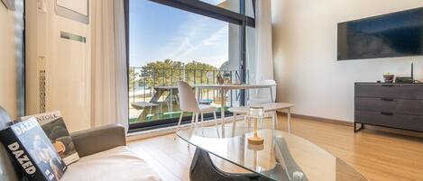 Una sala de estar moderna con una mesa de vidrio, un sofá, un televisor montado en la pared y un balcón con mobiliario exterior.