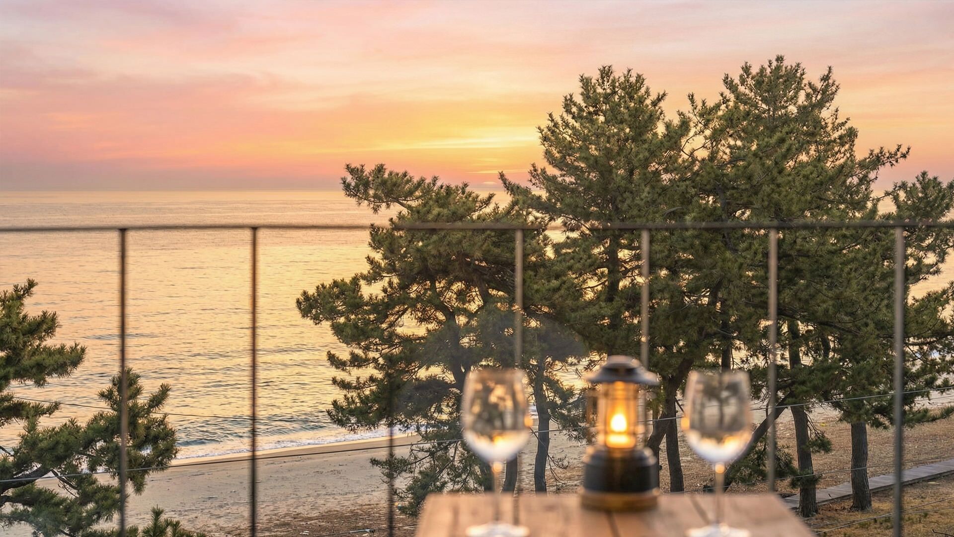 A wooden table with two wine glasses and a lantern, overlooking a beach with trees and a sunset.