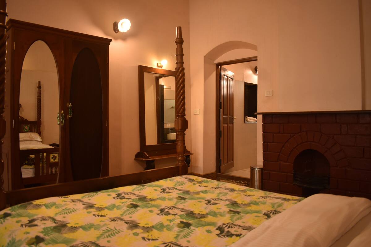 A bedroom with a large bed, a wooden wardrobe, a mirror, and a brick fireplace.