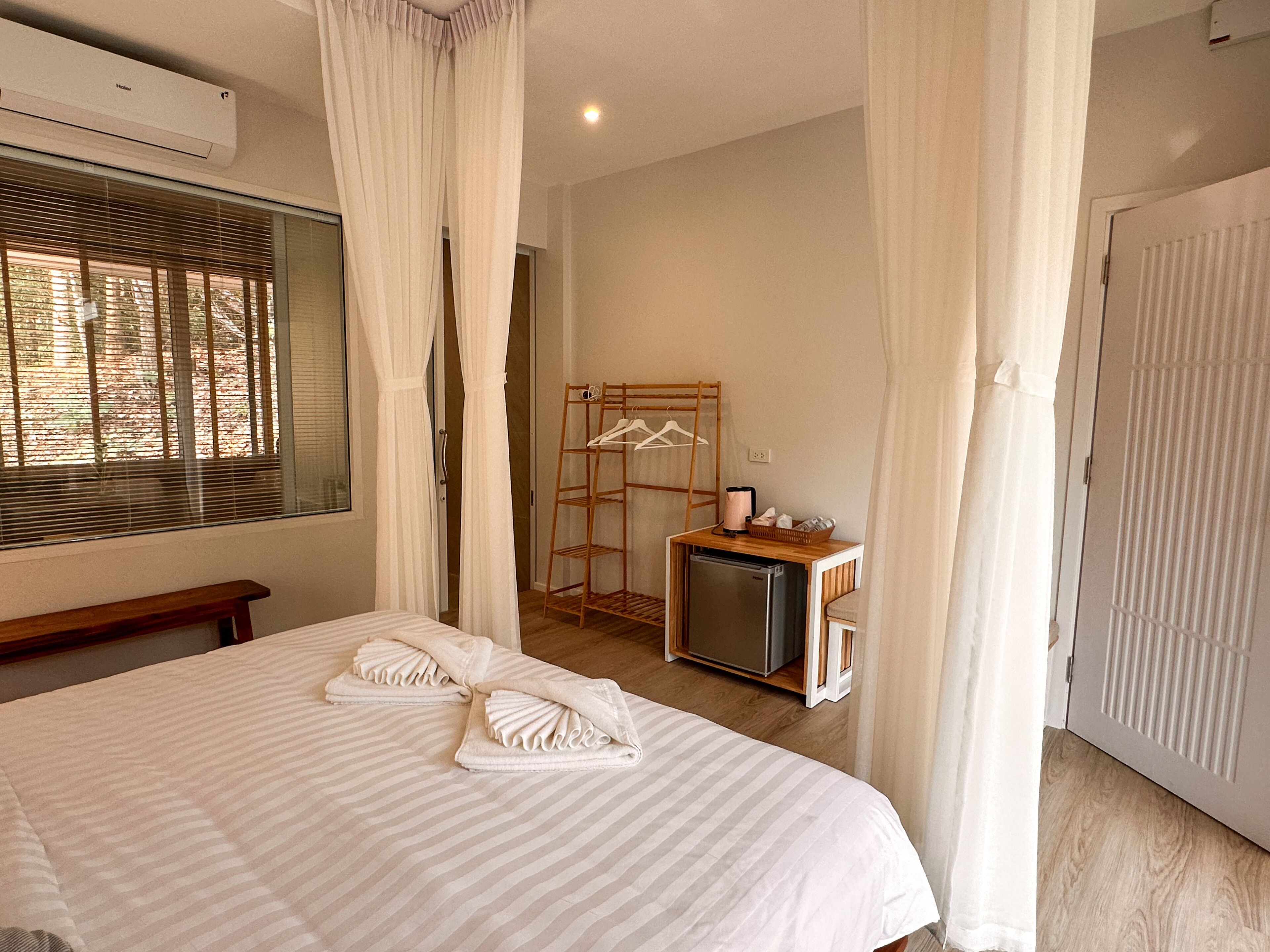 A hotel room with a bed, a small refrigerator, a wooden drying rack, and a window with blinds.