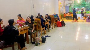A dining area with tables and chairs, a fan on the wall, and a view of a garden through the window. - Nam Anh Hotel (Phong Nha)