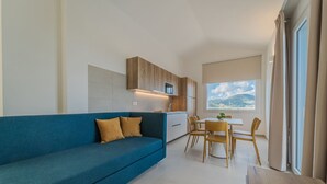 A modern living room with a blue sofa, wooden cabinets, a dining area with a table and chairs, and a large window with a view of hills. - RESIDENCE PADULELLA - 2 - RESIDENCE PADULELLA - ATTICO SX (Portoferraio)
