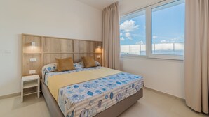A bedroom with a bed, bedside tables, a wooden headboard, a window with curtains, and a view of the sky.
