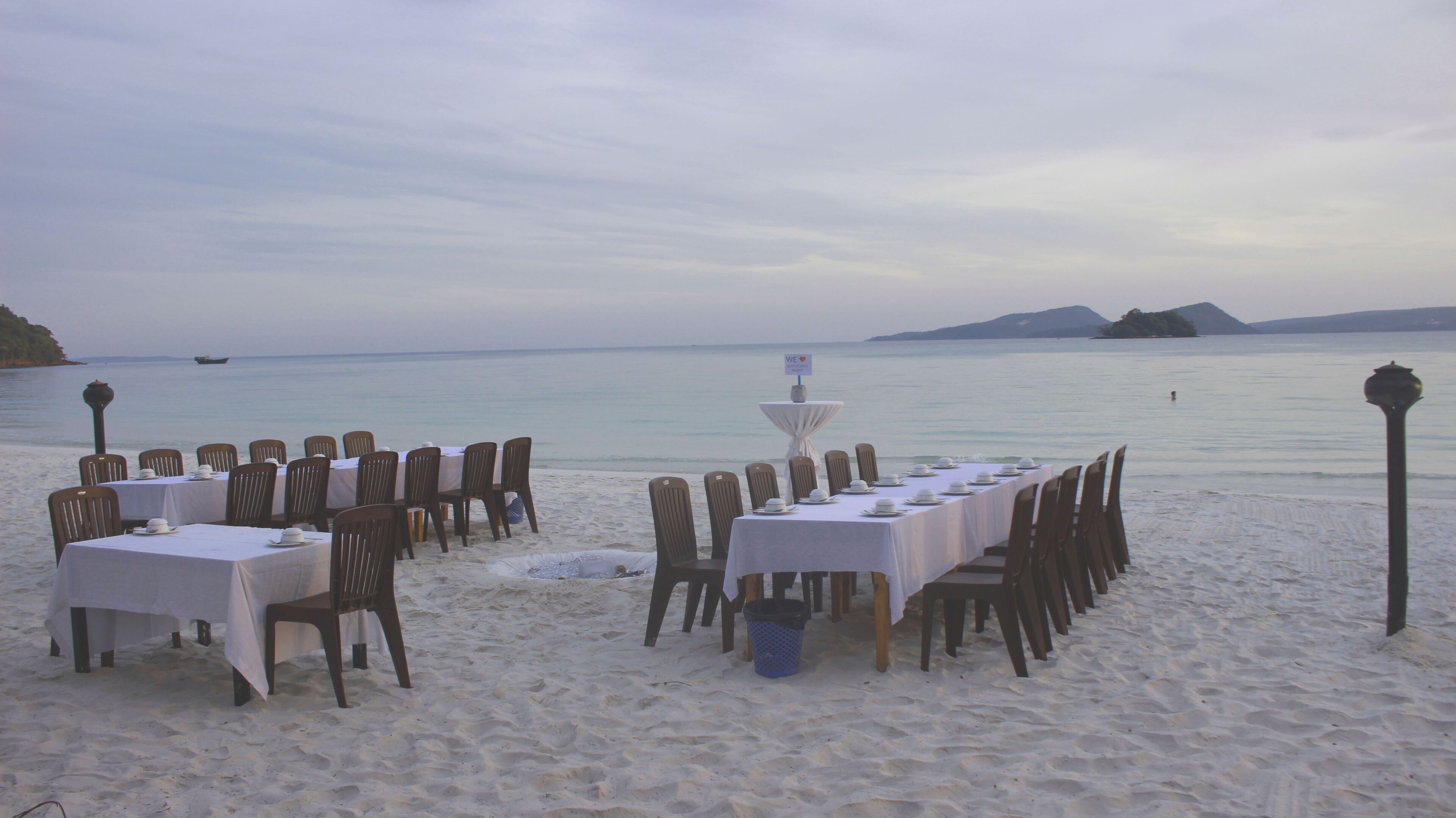 A beachside dining setup with tables and chairs arranged for a meal, overlooking a calm sea and distant islands.