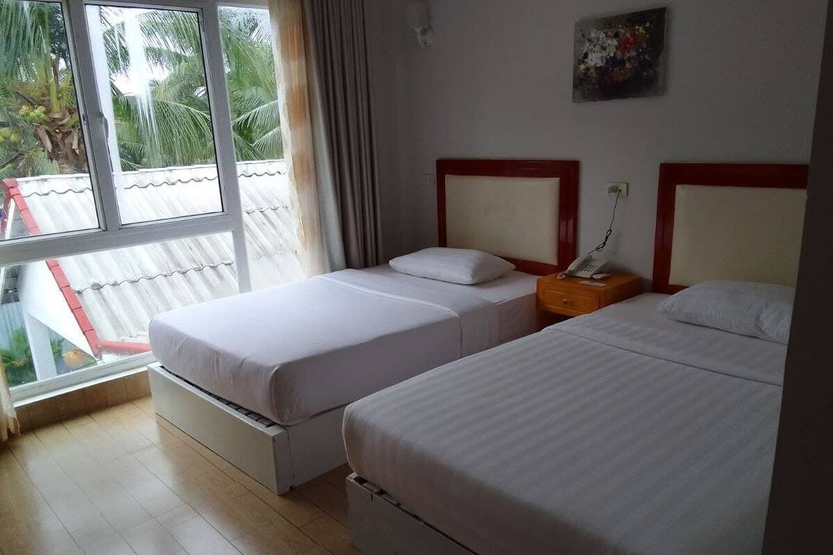 A hotel room with two beds, a window with a view of palm trees, and a small table with a lamp.