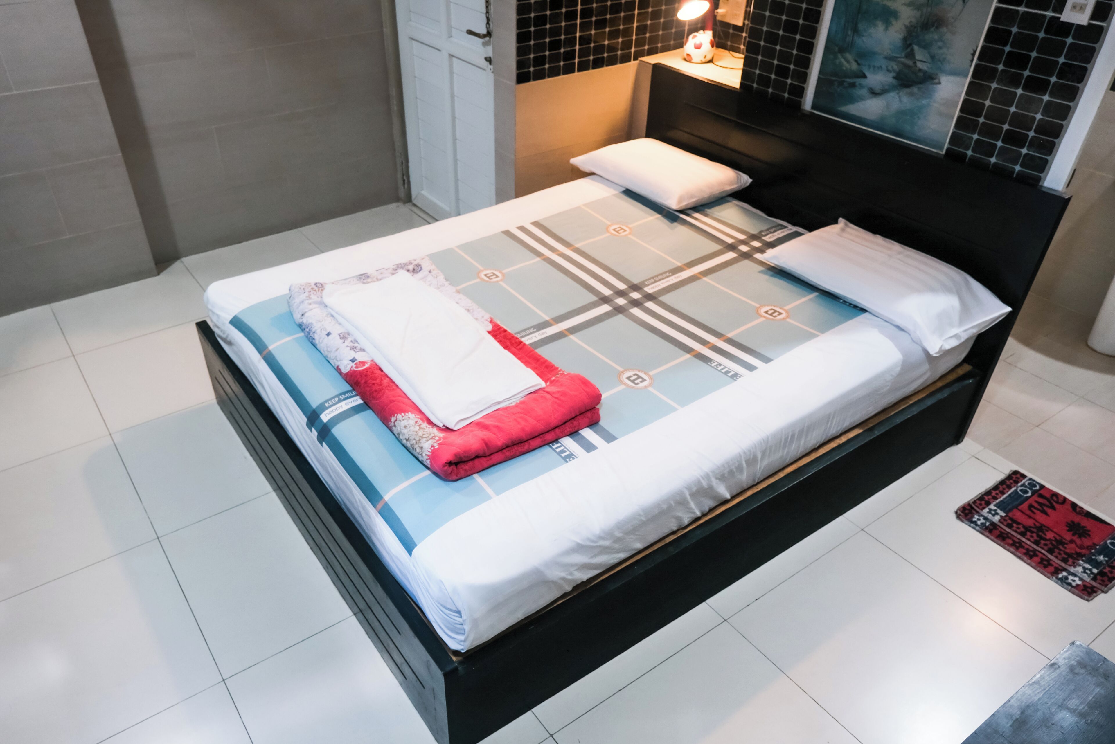 A neatly made bed with a plaid bedspread and a red blanket, a bedside table with a lamp, and a painting on the wall.