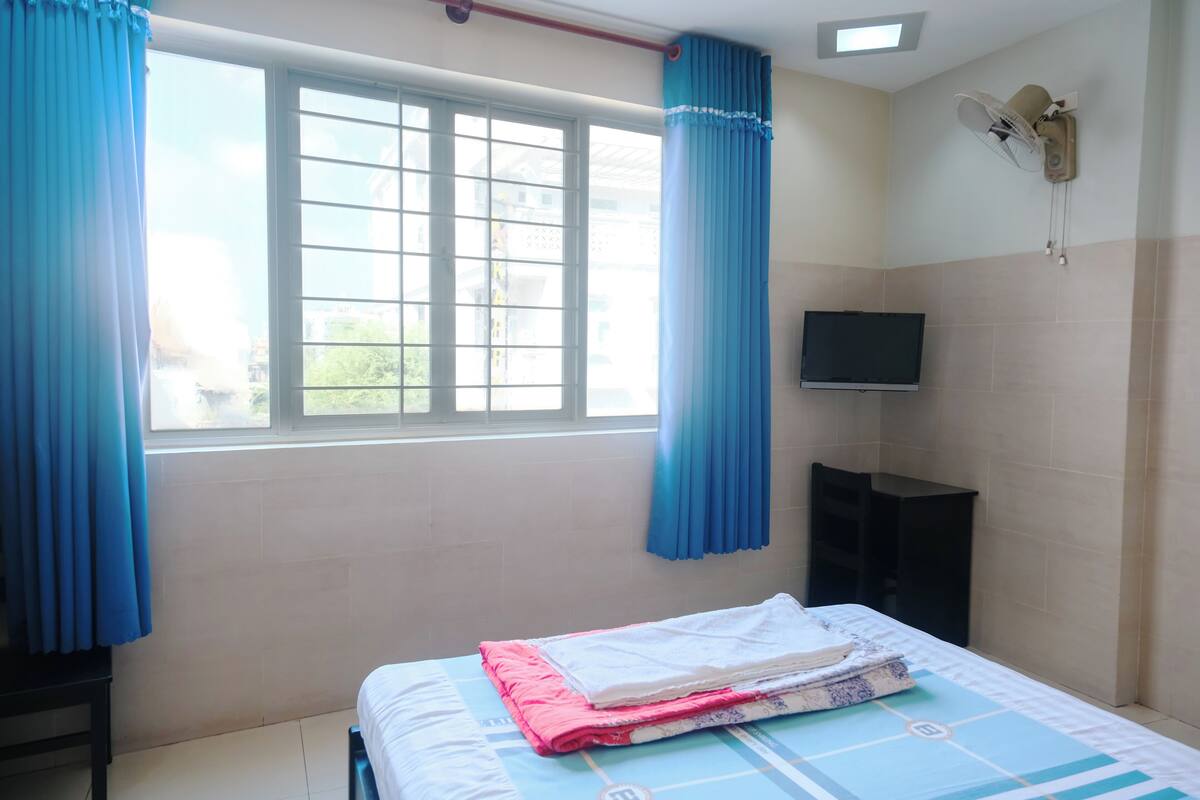 A hotel room with a bed, a TV, a wall-mounted fan, and a window with curtains.