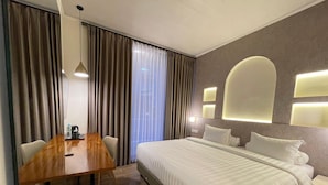 A hotel room with a wooden desk, a bed with white bedding, a large window with curtains, and wall-mounted lighting. - Azana Essence Sanggabuana Bogor (Bogor)