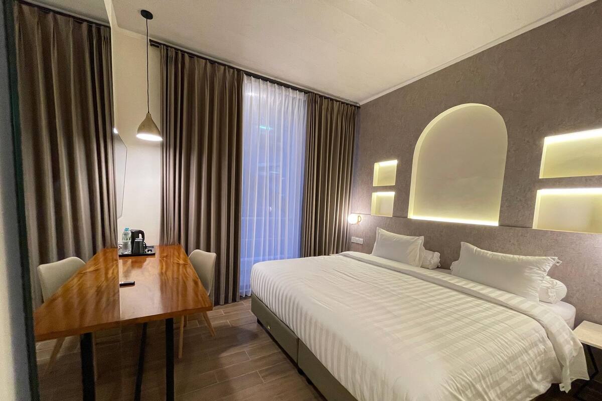 A hotel room with a wooden desk, a bed with white bedding, a large window with curtains, and wall-mounted lighting.