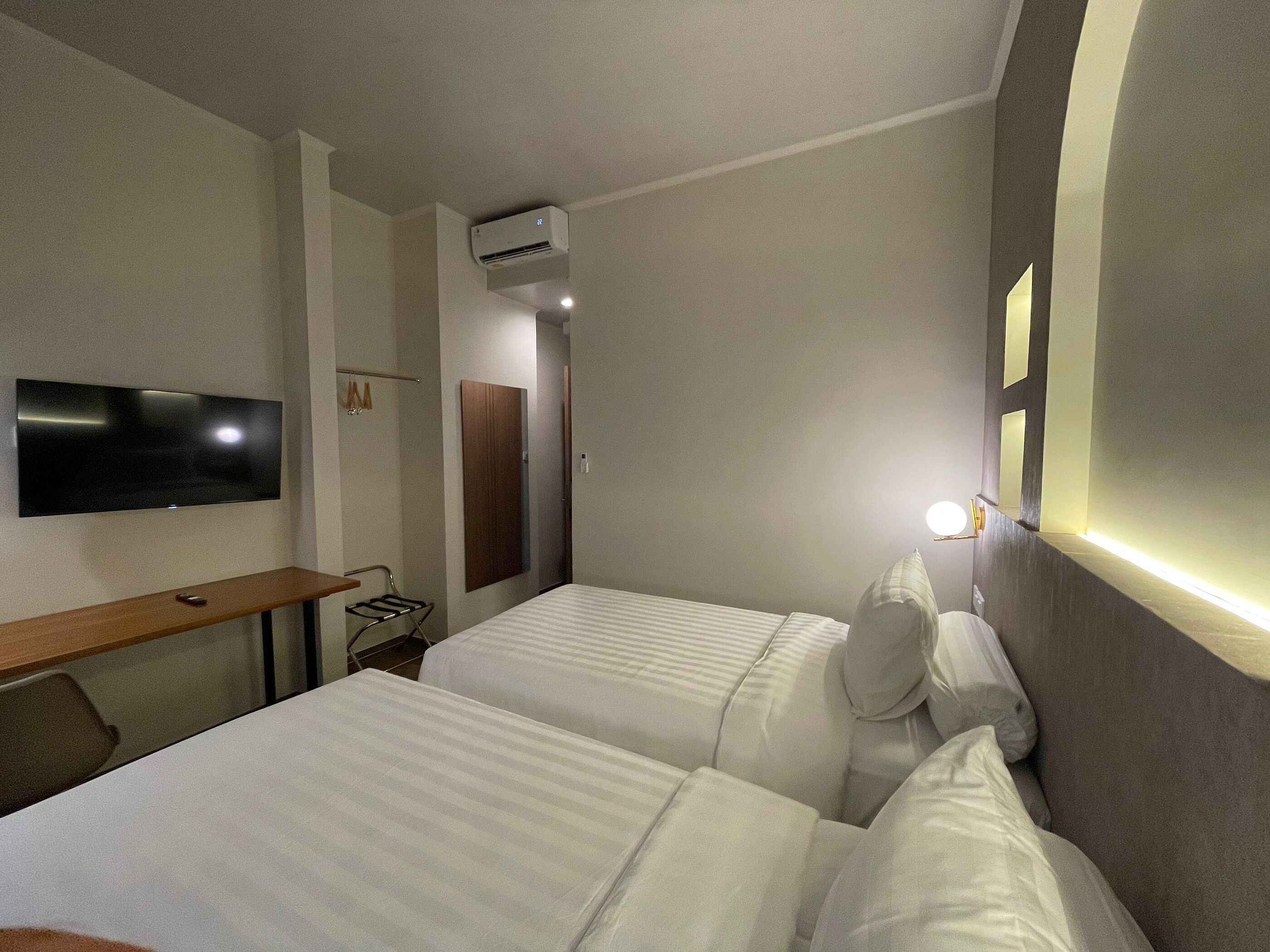 A hotel room with two beds, a flat-screen TV mounted on the wall, an air conditioning unit, a bedside table, and a lamp.