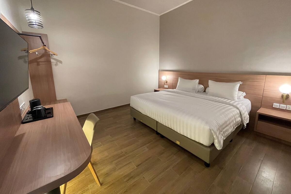A modern hotel room with a wooden bed, a desk with a chair, a flat-screen TV, and a wall-mounted lamp.