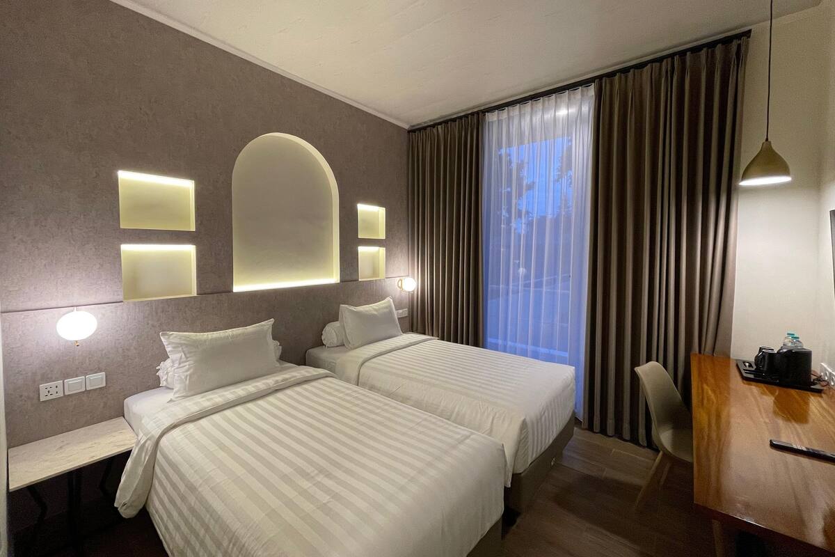 A hotel room with two beds, a desk, a chair, and a large window with curtains.