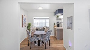 Un comedor con una mesa redonda y sillas estampadas, una cocina con encimera y armarios, y un ventanal con persianas.