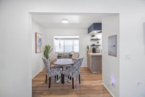 Zona pranzo con tavolo rotondo e sedie decorate, cucina con piano di lavoro e mobili, e una finestra con persiane.